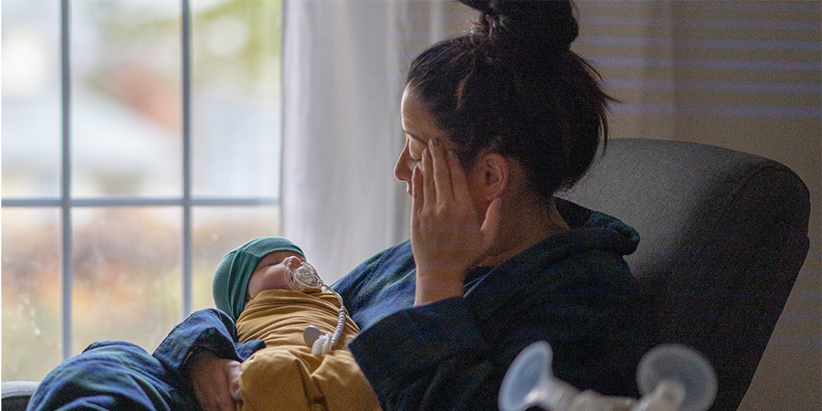 A new mother sitting on a rocking chair in her living room wearing a housecoat holding her newborn baby. There is a breast pump and nursing supplies around.